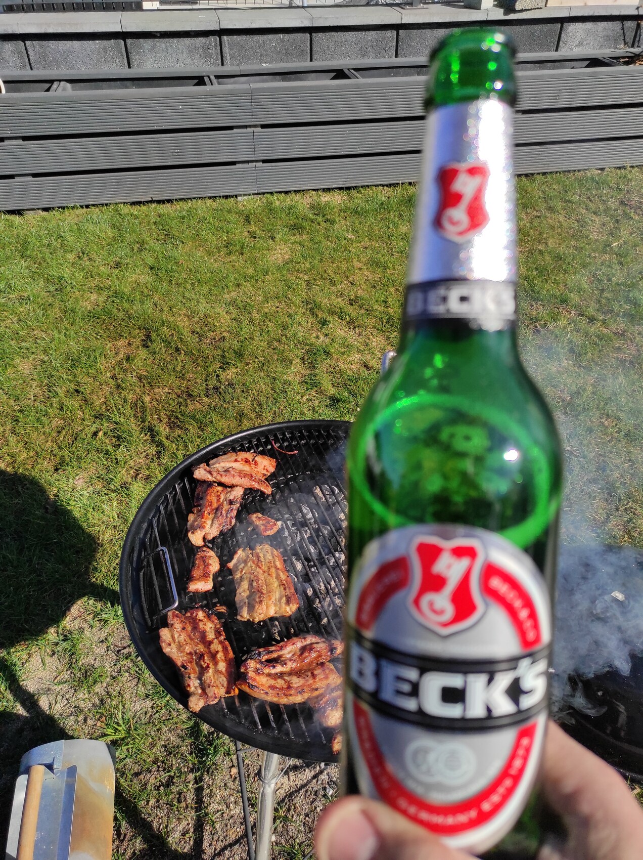 Beck's-Bierflasche im Vordergrund; dahinter grillt Fleisch auf einem runden Grill im Gras.