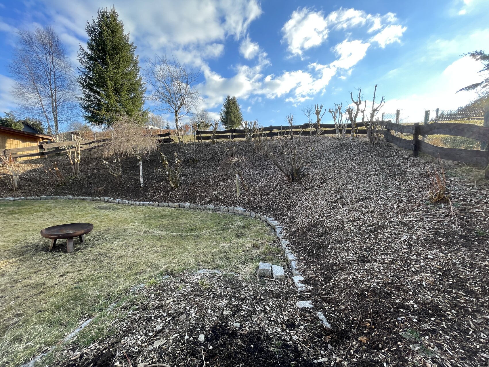 Garten auf Hang mit Rindenmulch, kahlen Sträuchern, Holzzaun; Feuerstelle im Gras.