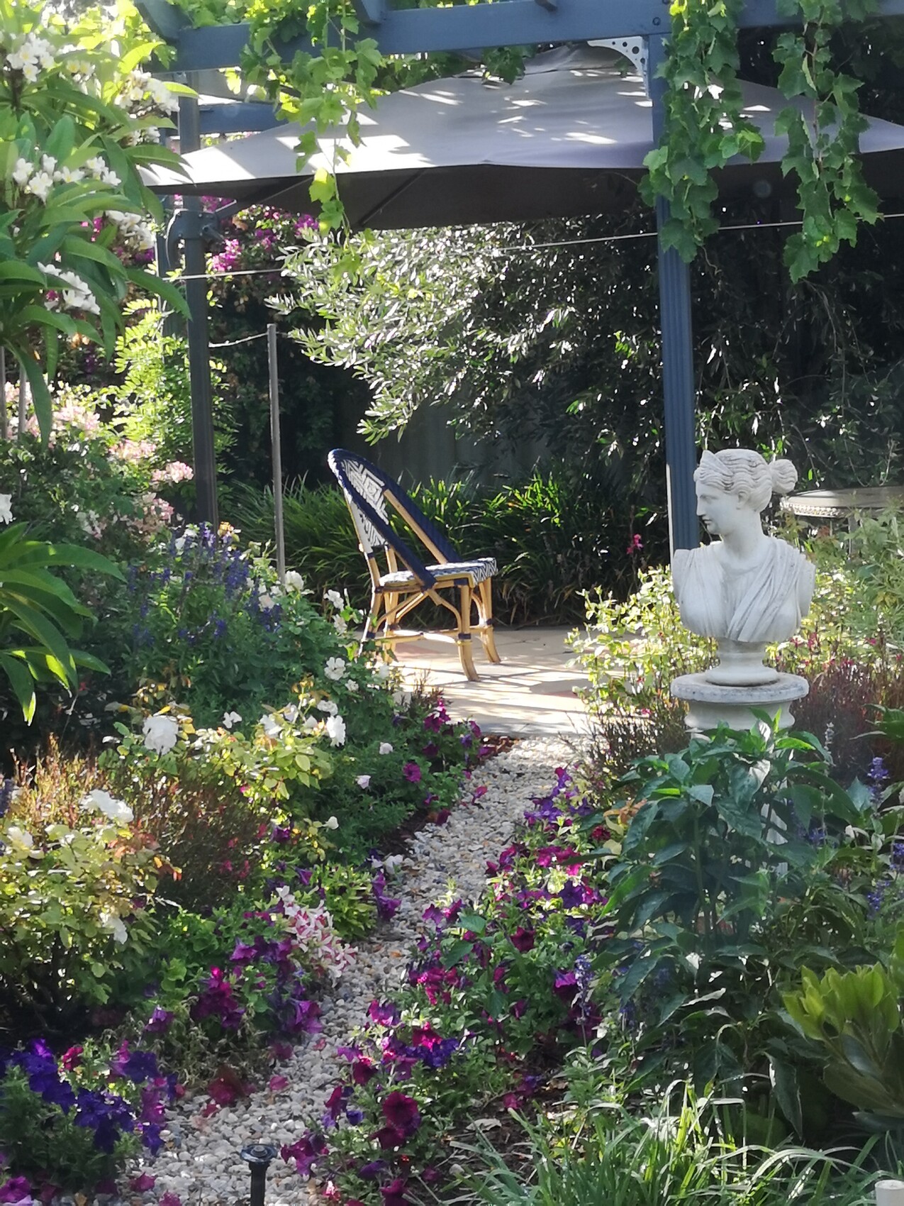 Gartenlandschaft mit Kiesweg, bunten Blumen, Statue und Sitzplatz unter einer Pergola im Garten.