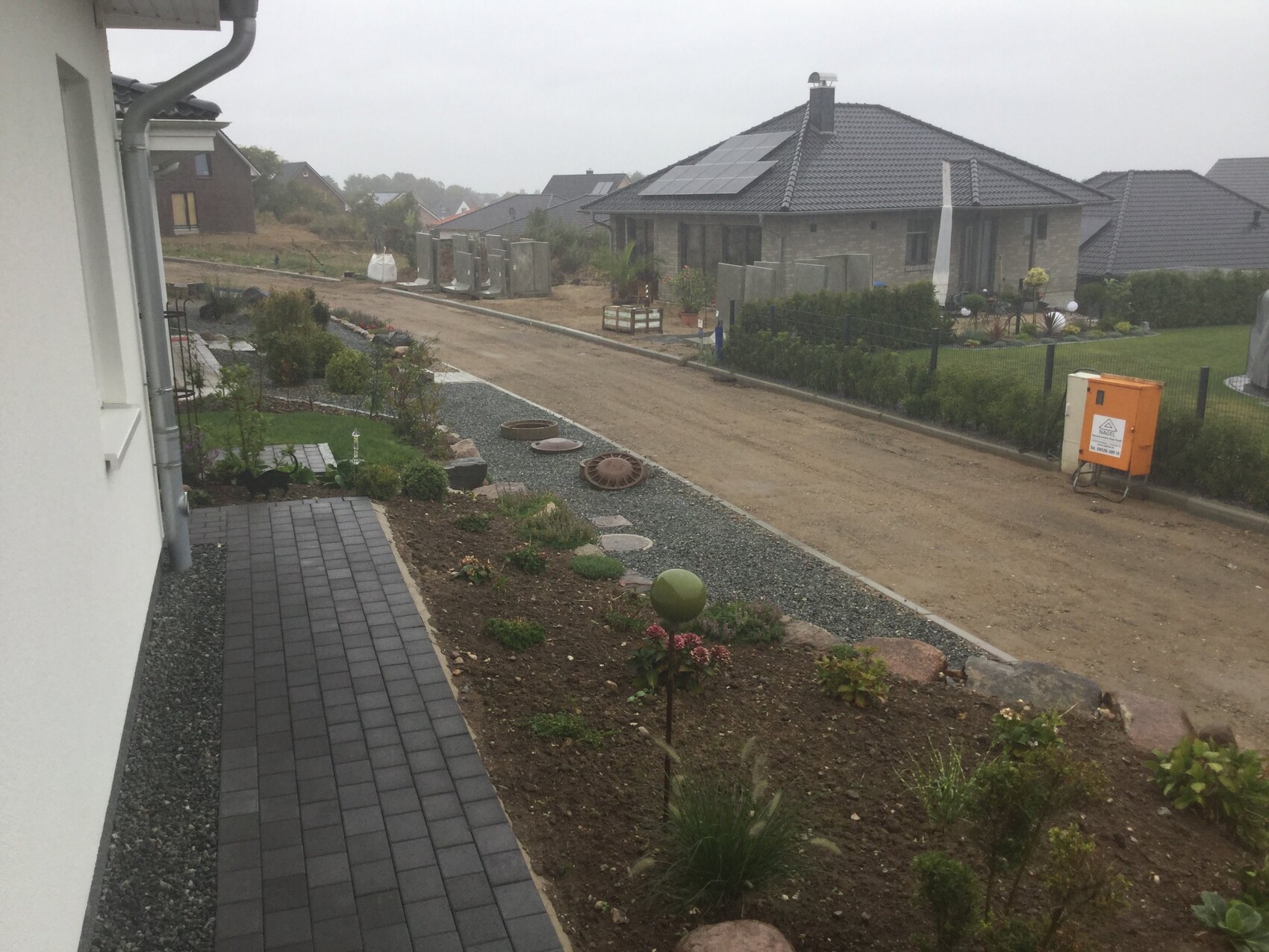 Vorderansicht eines Neubauviertels: Gehweg neben Garten, unbefestigte Straße, orange Verteilerbox.