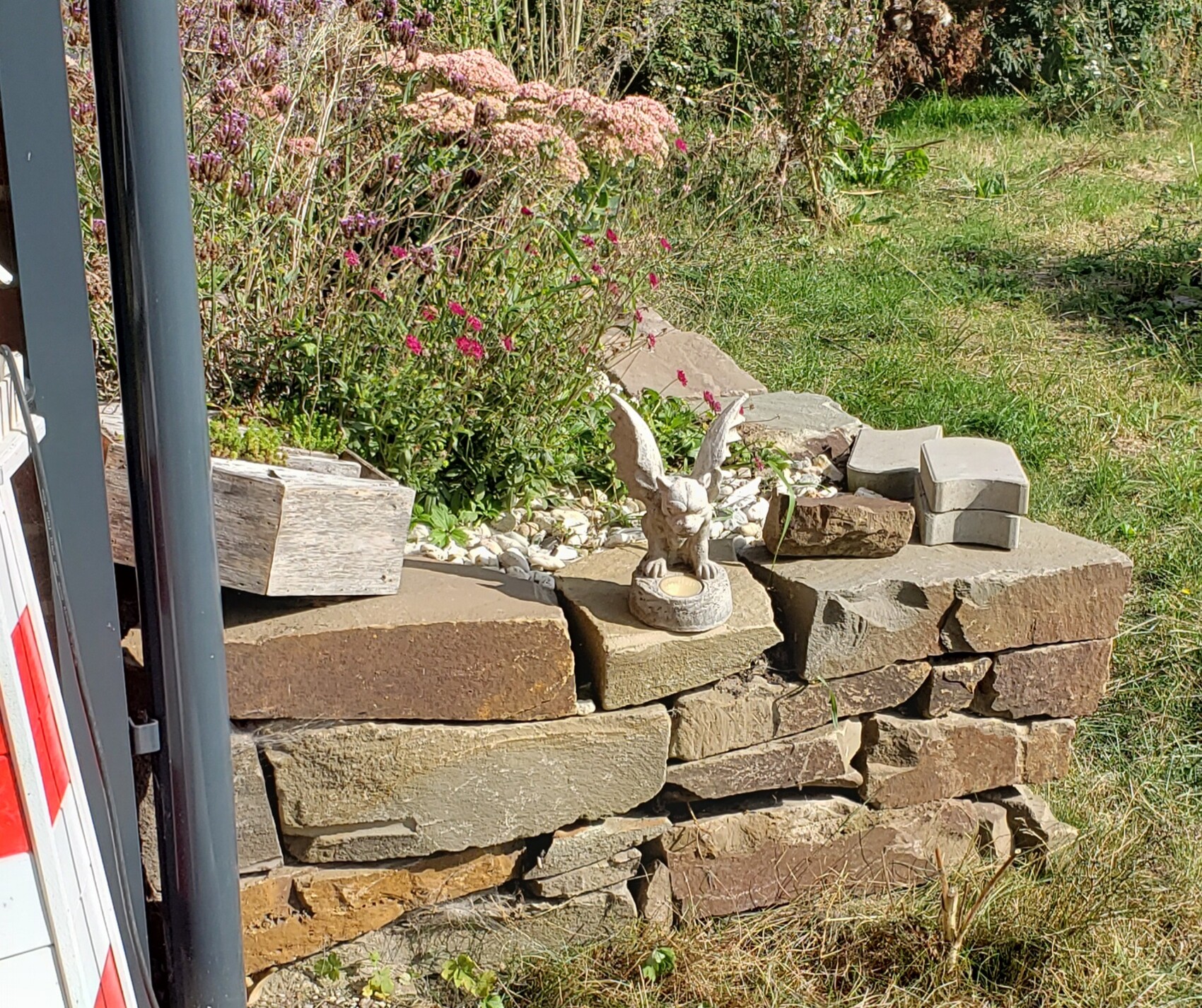 Kleine Engelfigur mit Flügeln sitzt auf einer Steinmauer im Garten, neben Pflanzkästen und Blumen.