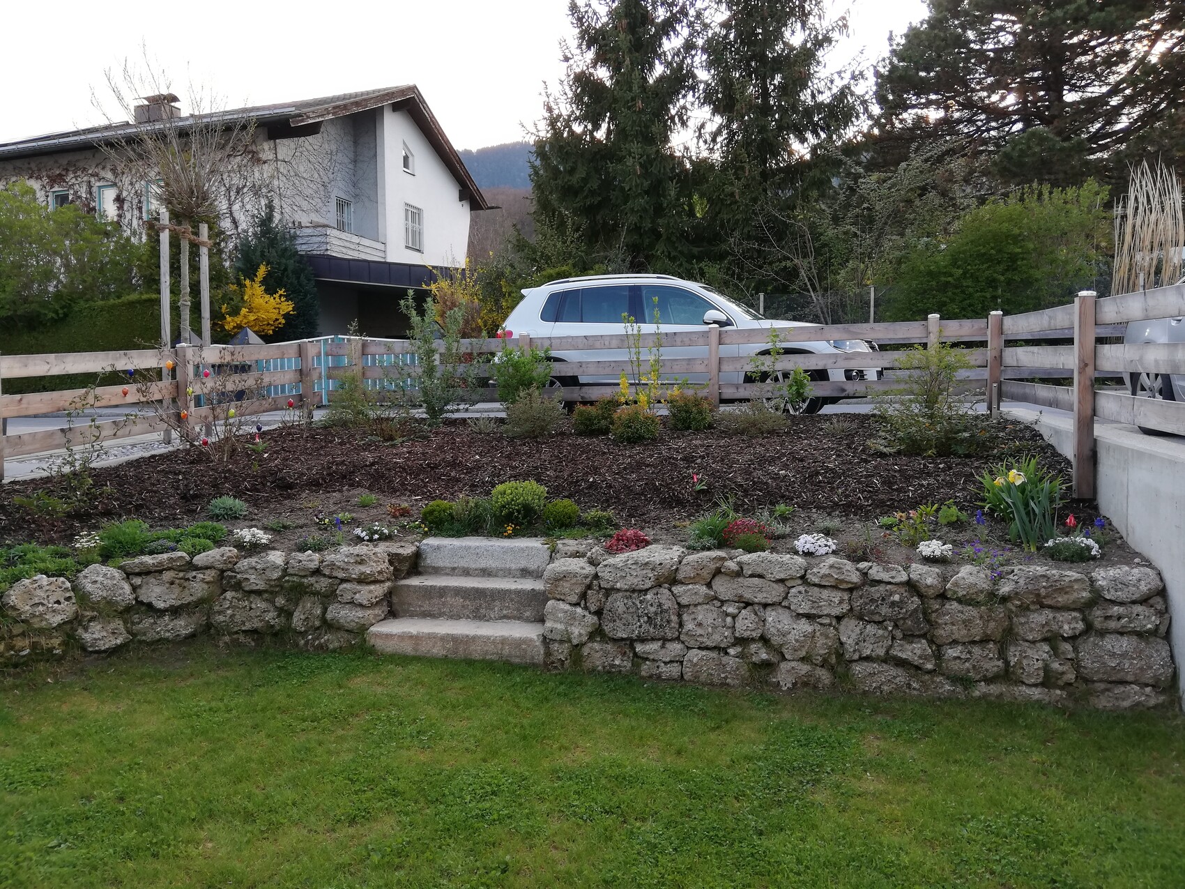 Garten mit Steinmauer, Treppenstufen und Blumenbeeten; Auto im Hintergrund