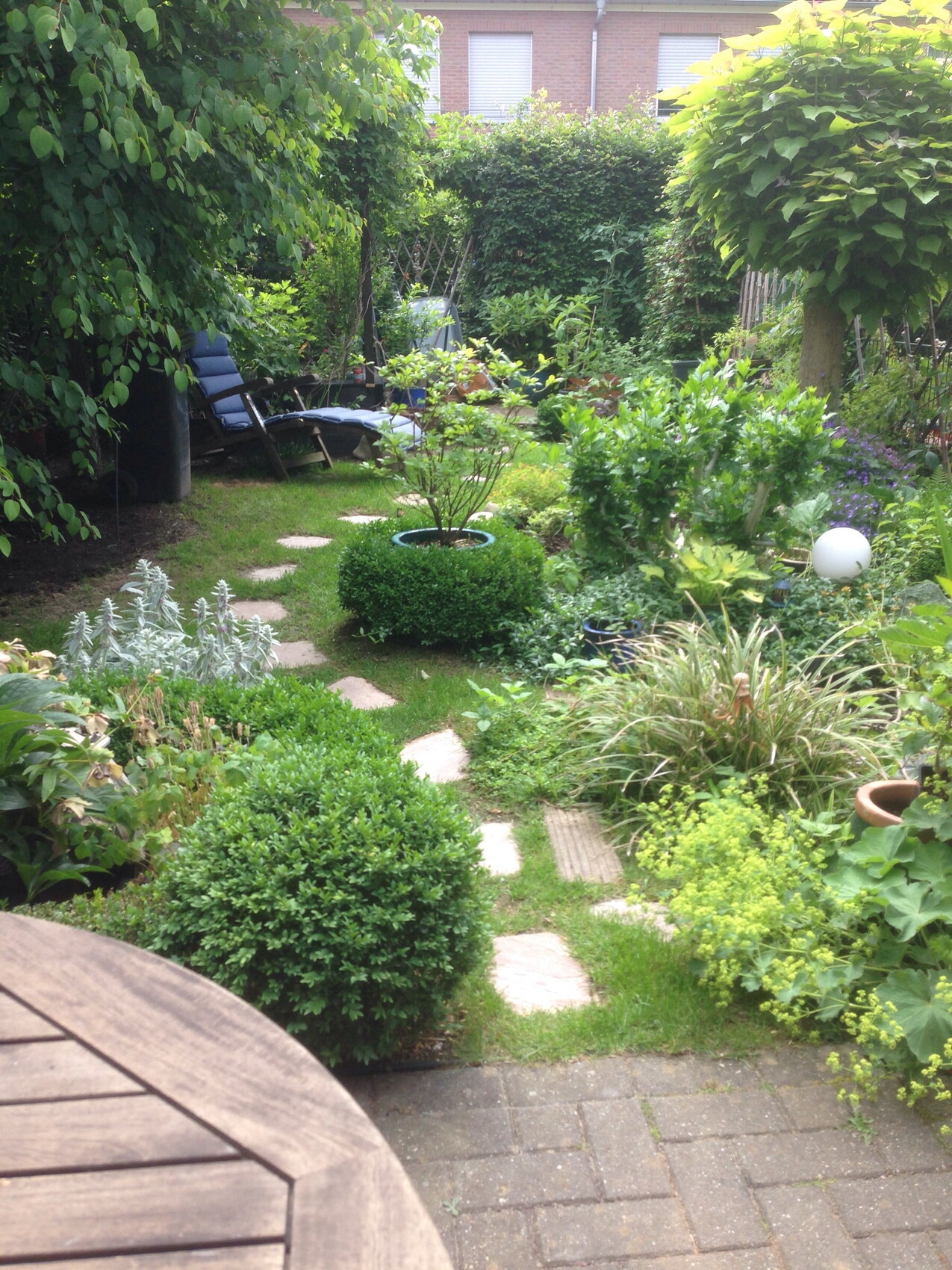 Garten mit Steinpfad, Beeten und rundem Holztisch im Vordergrund