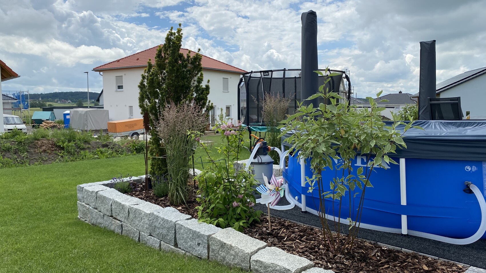 Garten mit blauem Above-Ground-Pool, Trampolin, Stein-Beet, grünem Rasen und Haus im Hintergrund.