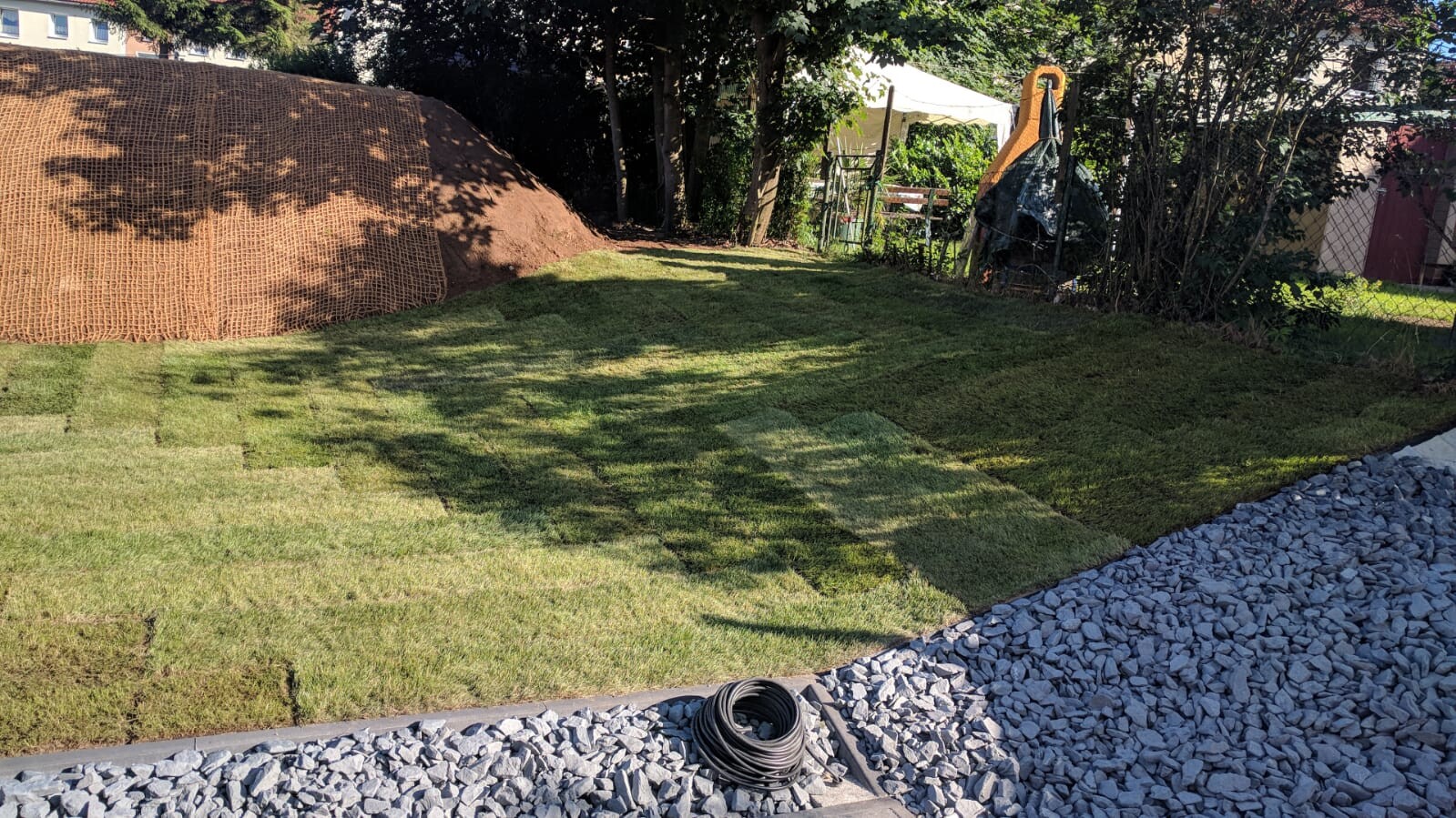 Garten mit neuem Rollrasen; links Erdhügel mit Netzabdeckung, vorn Kiesweg und Kabelrolle.