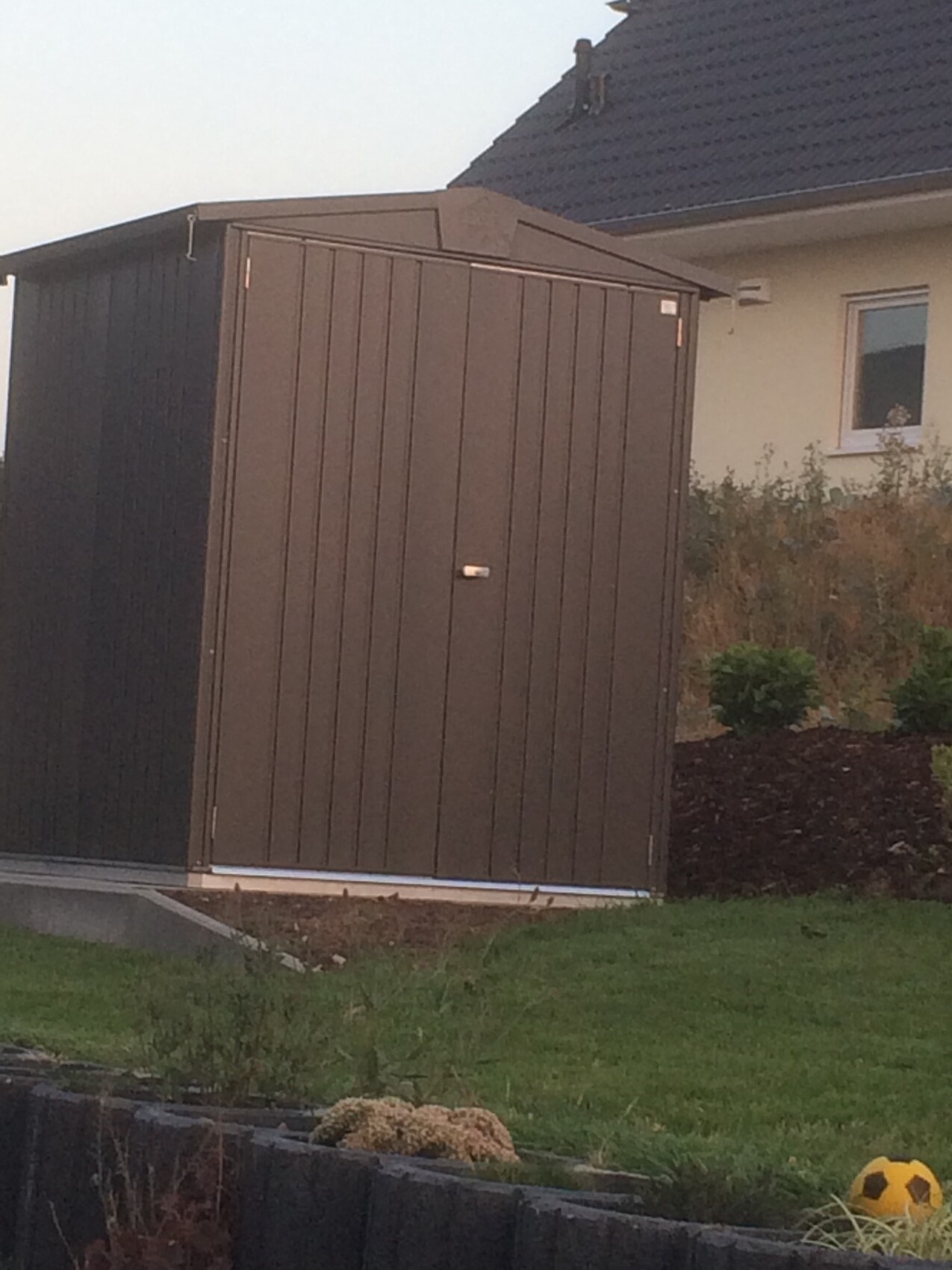 Braunes Gartenhaus/Schuppen vor Wohnhaus; Rasen, Sträucher, und ein gelber Fußball auf der Wiese.