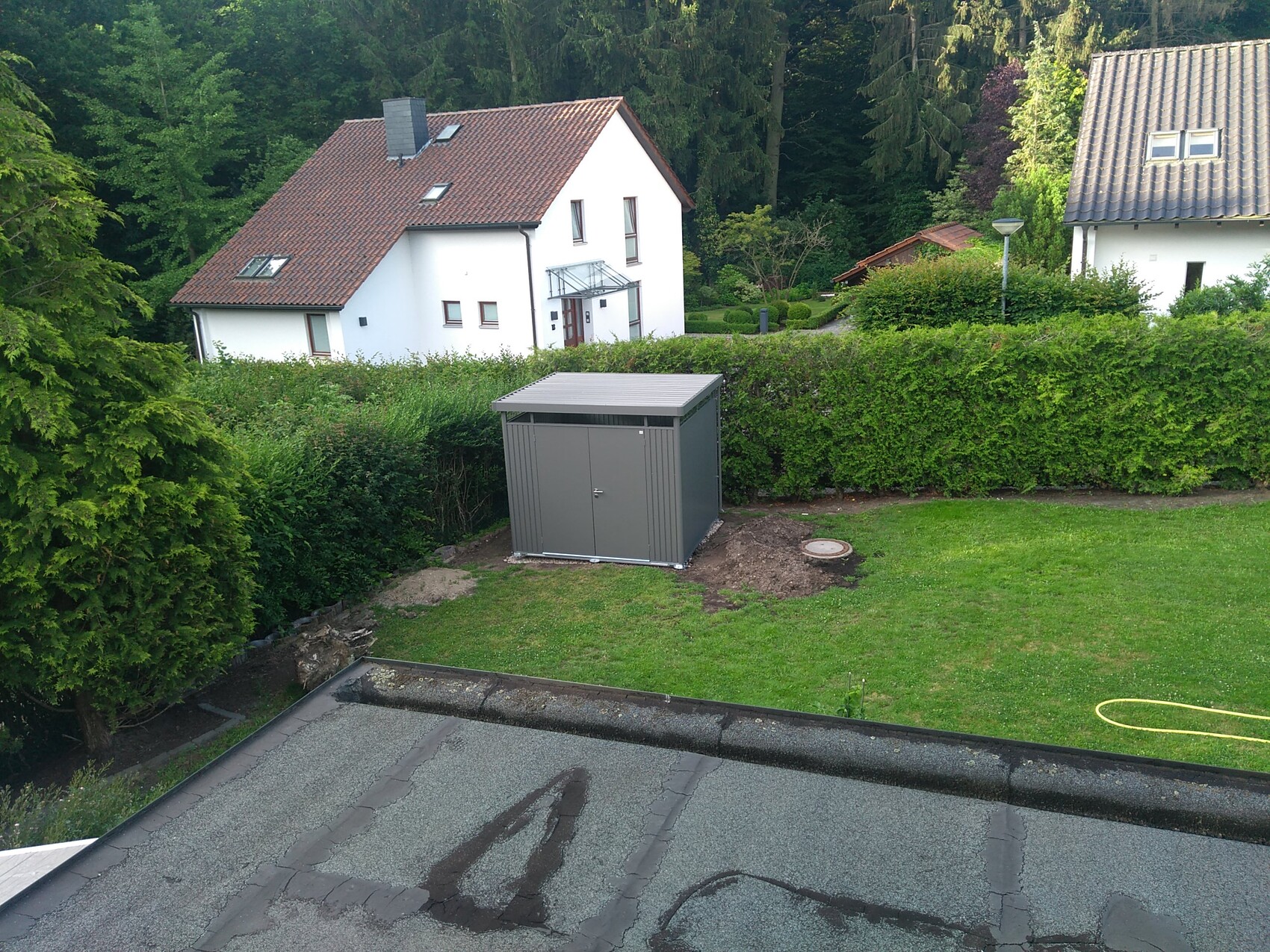 Gartenansicht mit grauem Metallschuppen vor grüner Hecke; weißes Haus dahinter.