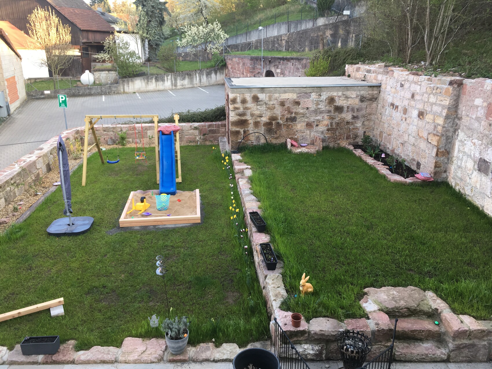 Hinterhof-Spielplatz: Sandkasten, blaue Rutsche, Schaukel, Steinmauer und Rasen.