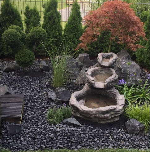 Garten mit mehrstufigem Steinbrunnen, Kiesboden, Zierpflanzen und Hintergrundhecke