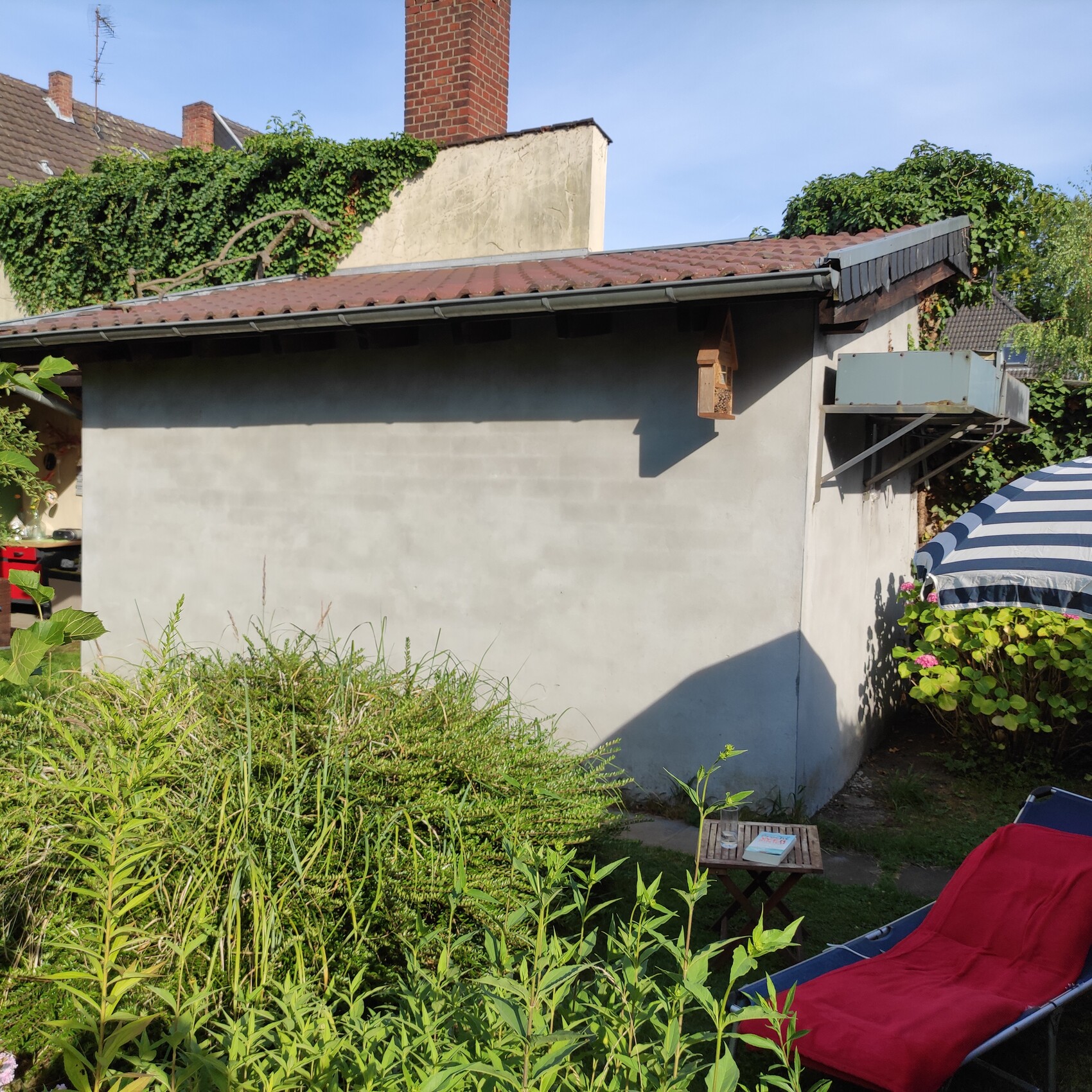 Kleines Gartenhaus mit rotem Ziegeldach, Gartenpflanzen, Lounge-Stuhl und Sonnenschirm