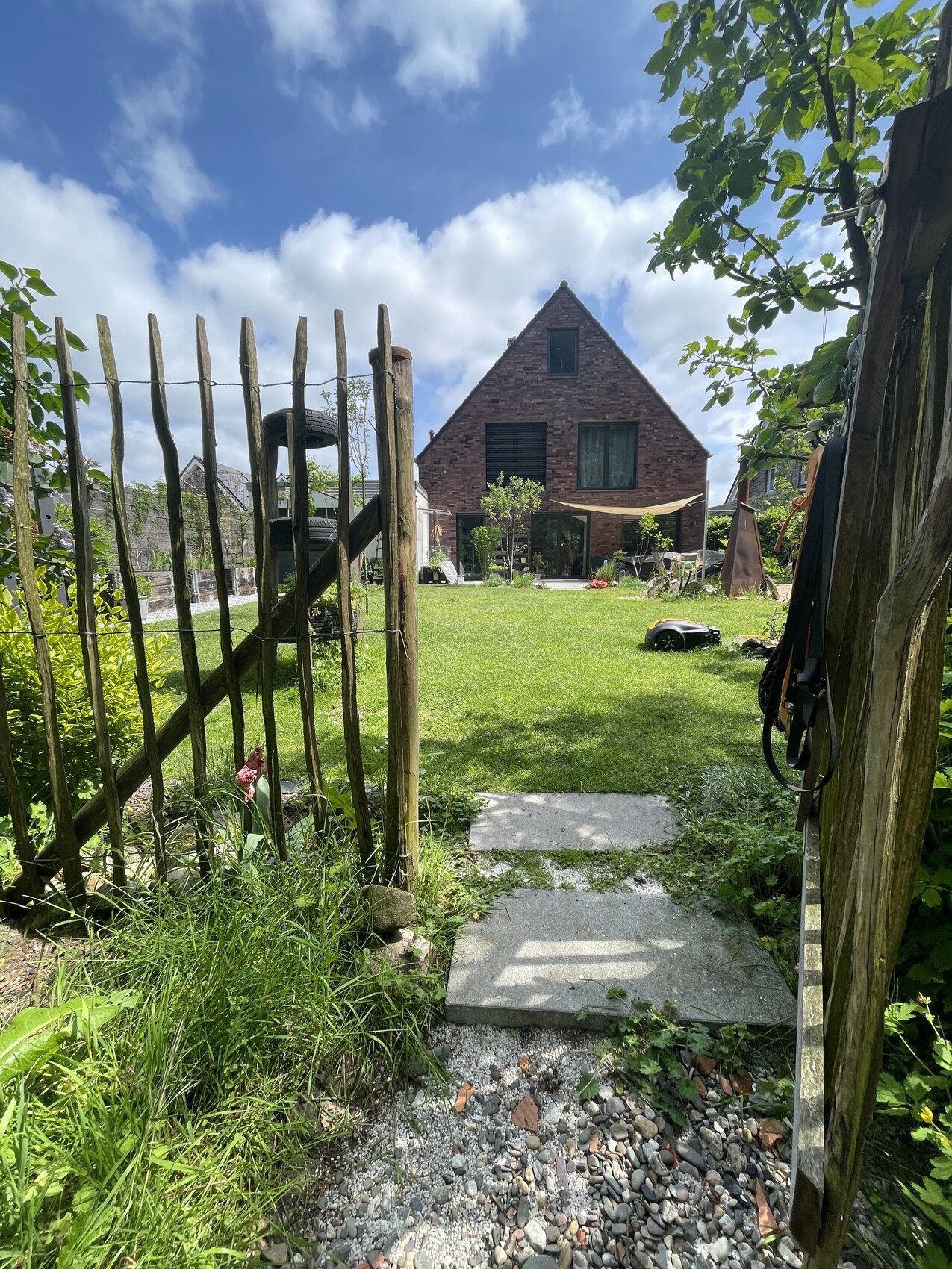 Garten mit Holzzaun, rotem Backsteinhaus, grüne Wiese, Kies- und Steinpfad.
