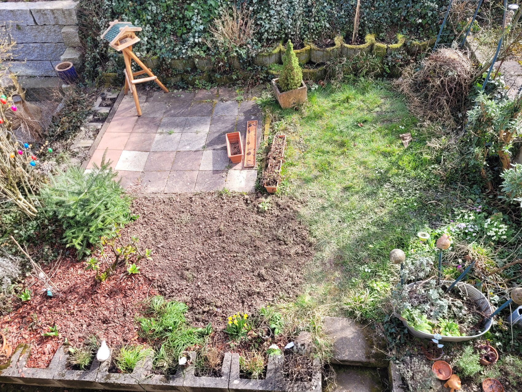 Terrasse im Garten mit gepflastertem Bereich, Vogelhäuschenständer, Pflanzkübeln, Beete und Rasen.