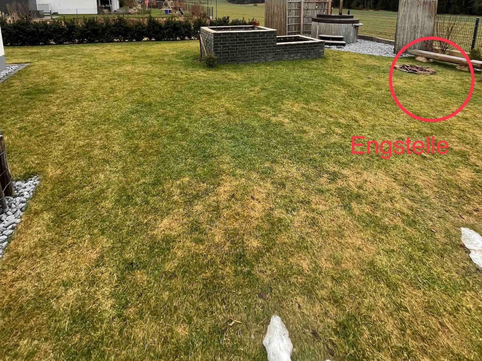 Garten mit grünem Rasen, zentrale Stein-Feuerstelle und Brennholzstapel neben einer Bank am Zaun.