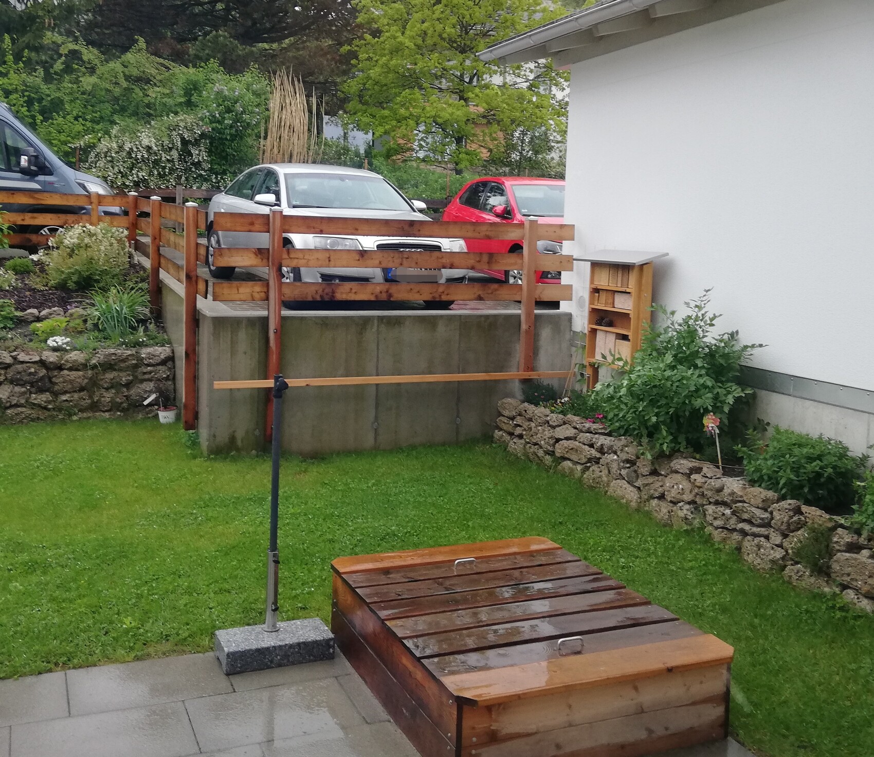 Garten mit Holzzaun und erhöhter Parkplatzfläche; silbernes und rotes Auto stehen am Zaun.