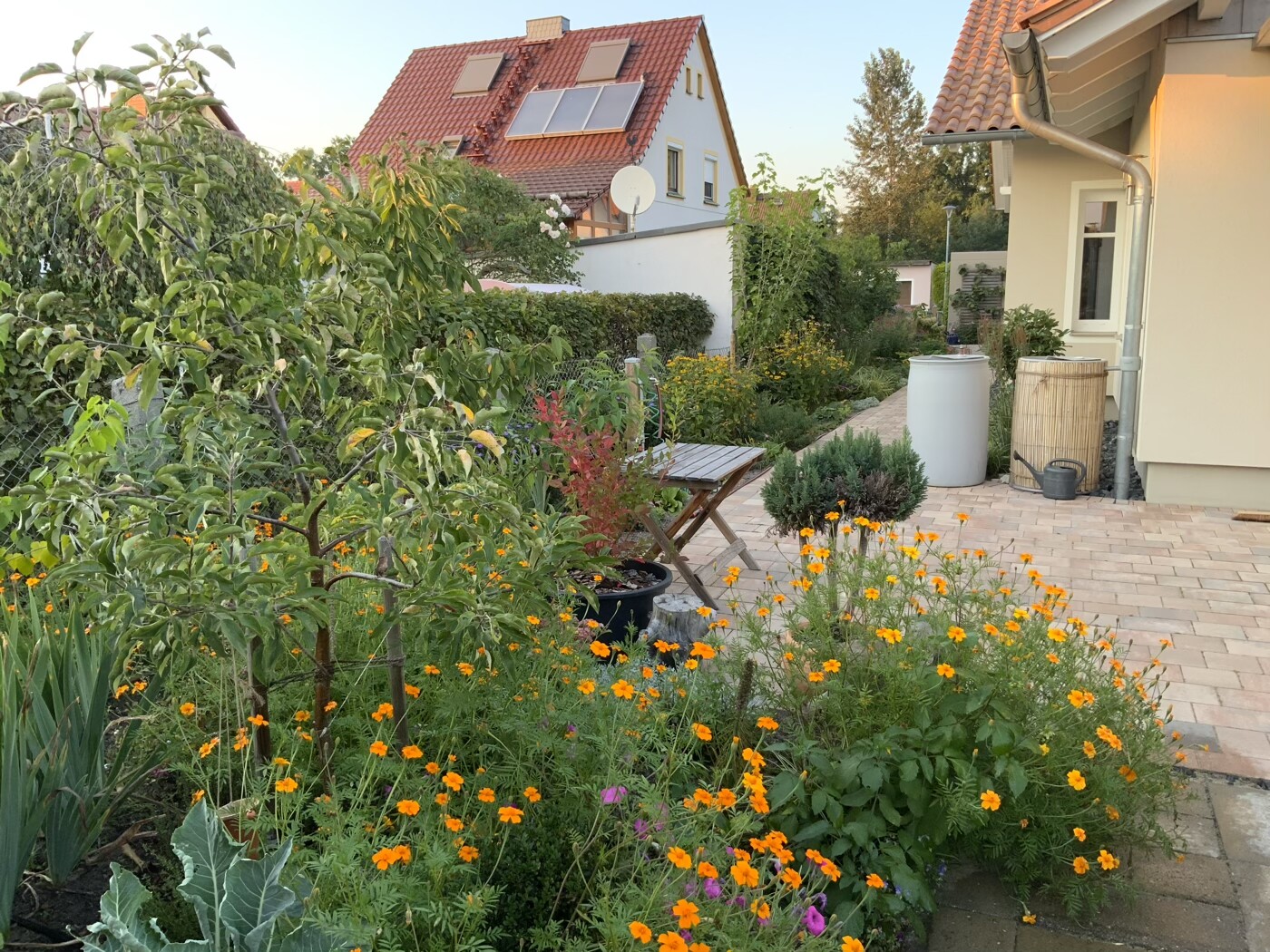 Gartenbereich vor Haus mit rotem Ziegeldach, Solarpaneele, blühende Blumen, Holztisch und Stühle.