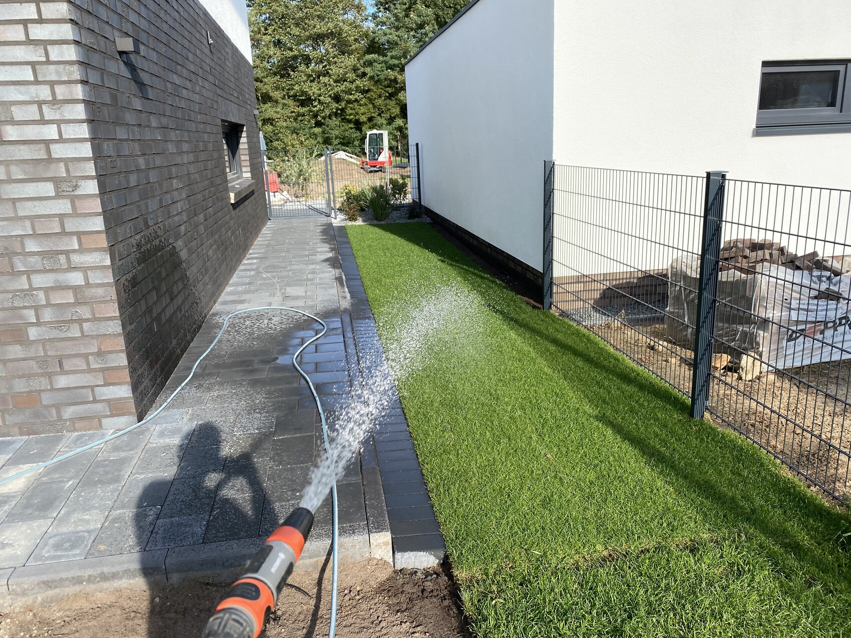 Gartenweg zwischen dunklem Ziegelhaus links und weißem Gebäude rechts; Schlauch sprüht Wasser auf Gras.
