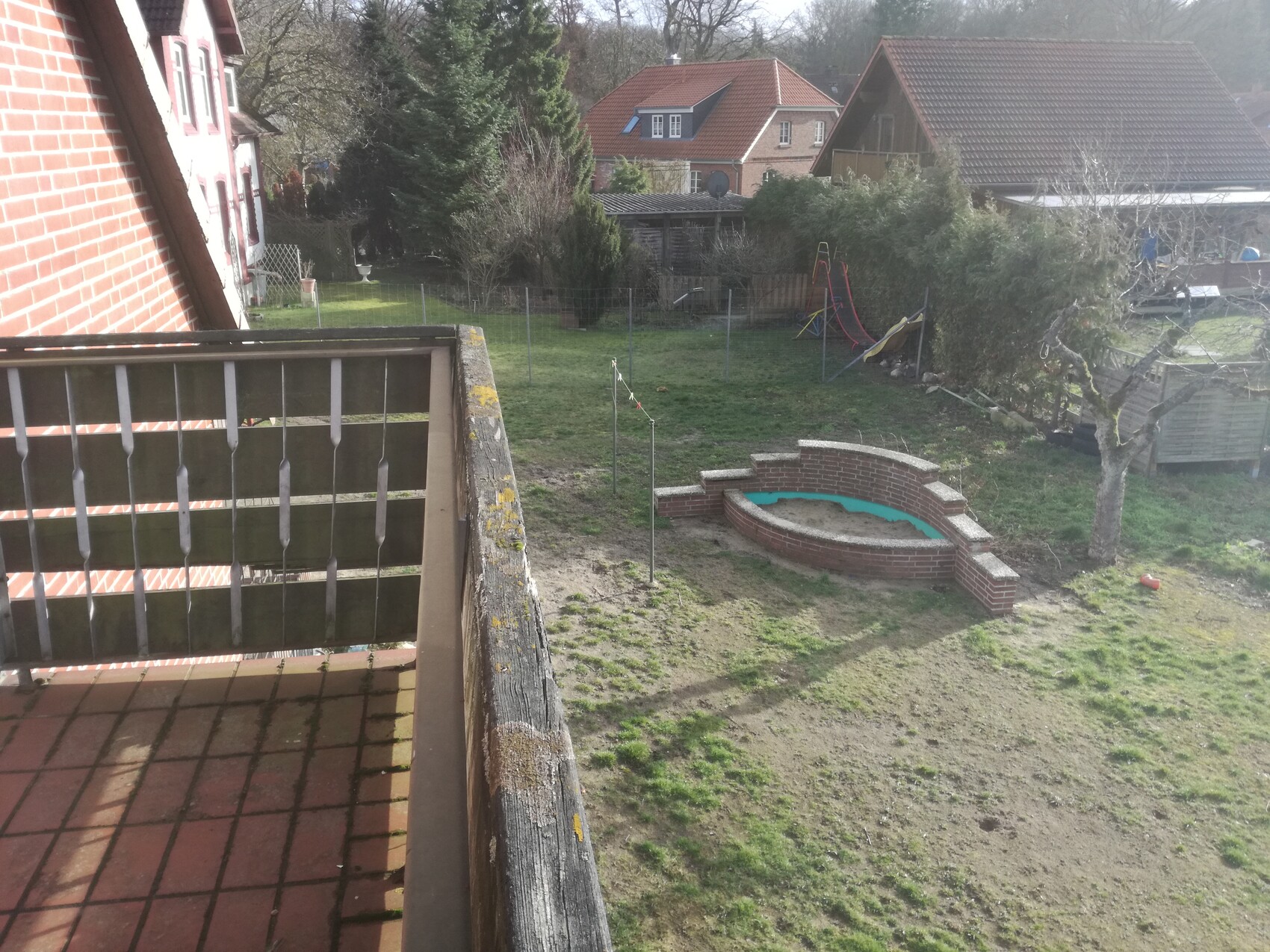 Blick vom Balkon in den Garten: Sandkasten aus Backstein, Spielgerät, Bäume, Häuser im Hintergrund.
