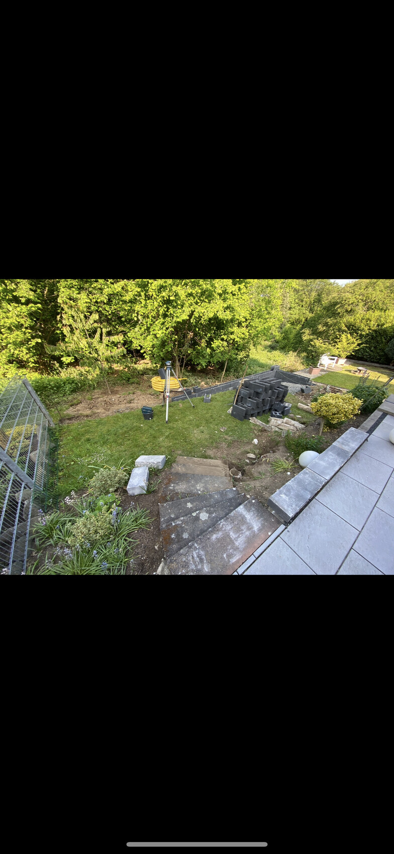 Gartenbereich hinter Terrasse: Steinstufen, Blockstapel, Rasen und Bäume im Hintergrund.