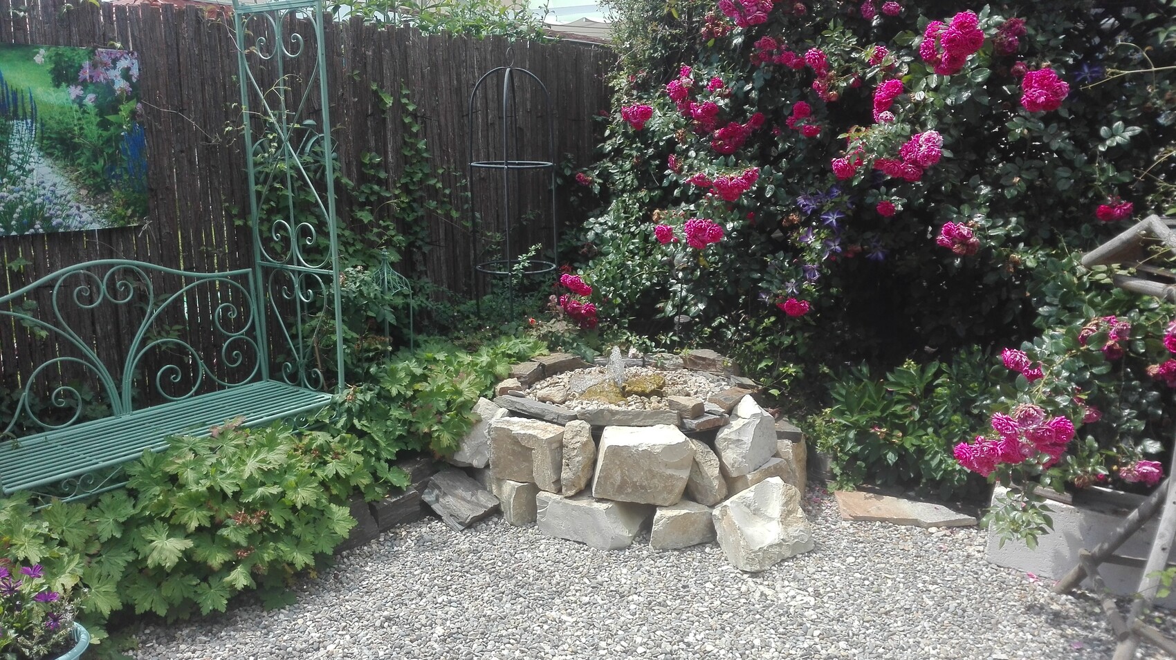 Garten mit grüner Schmiedeeisenbank, pinken Rosen und Steinring-Wasserbrunnen auf Kies.