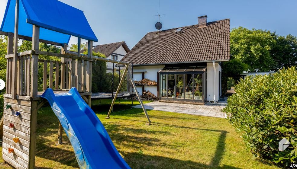 Garten mit Spielturm und blauer Rutsche, angrenzende Terrasse am Haus mit Glasfront und Pflanzen.