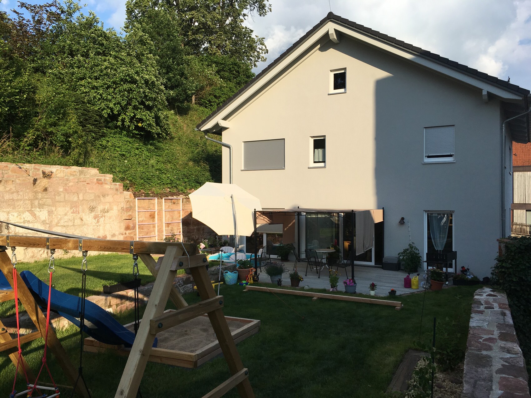 Zweigeschossiges Haus mit Terrasse, Sonnenschirm, Gartenmöbeln, Spielgeräten und Pflanzen.