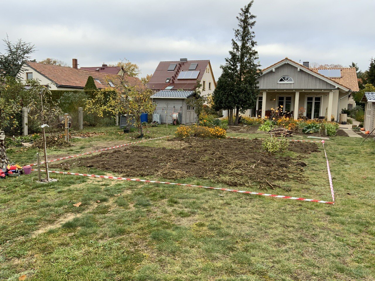 Vordergarten mit Beeten, Bäumen, Haus im Hintergrund und rotem Absperrband
