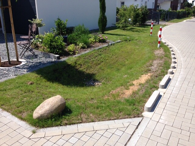 Grüner Gartenstreifen neben gepflasterter Straße, großer Stein im Gras, Sitzgruppe am Rand