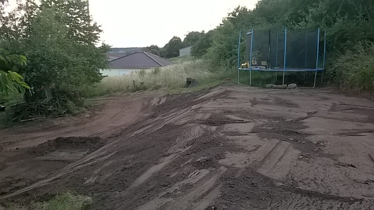 Gartenbereich mit offener Erde und Trampolin, umliegende Bäume und Häuser im Hintergrund.