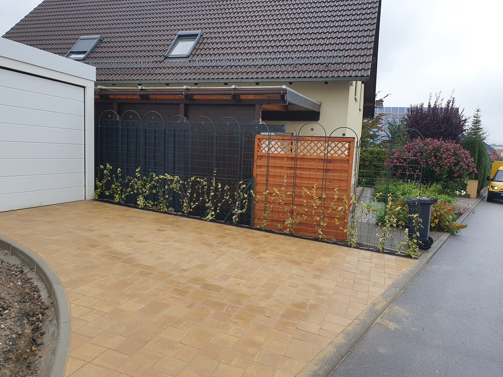 Frontansicht einer gepflasterten Einfahrt, Gartenzaun, Kletterpflanzen, Holzpaneel und Mülltonne.