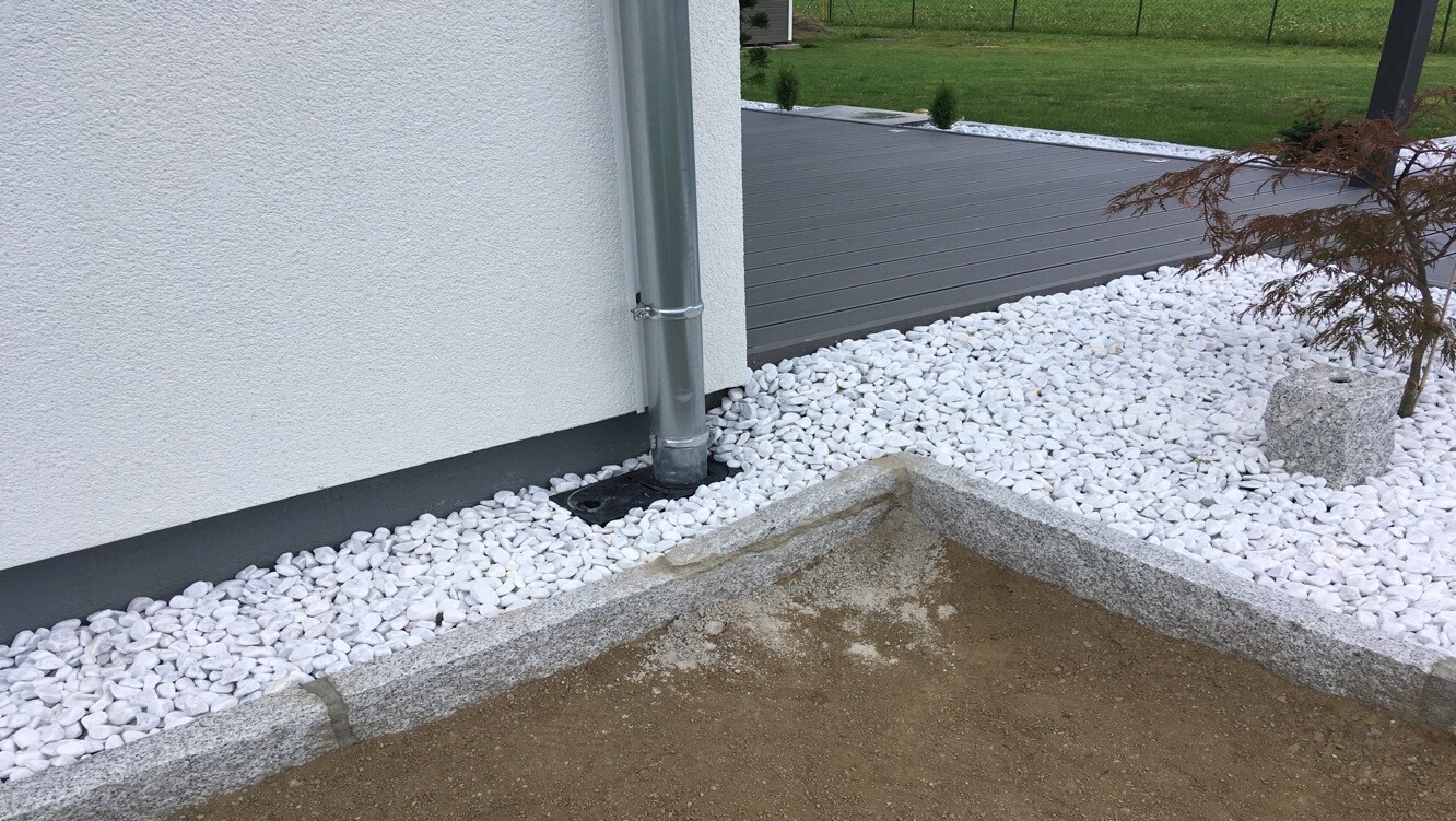 Außenwand eines Hauses mit Metall-Regenrohr, weiße Kiesel um die Wand und Baum im Garten