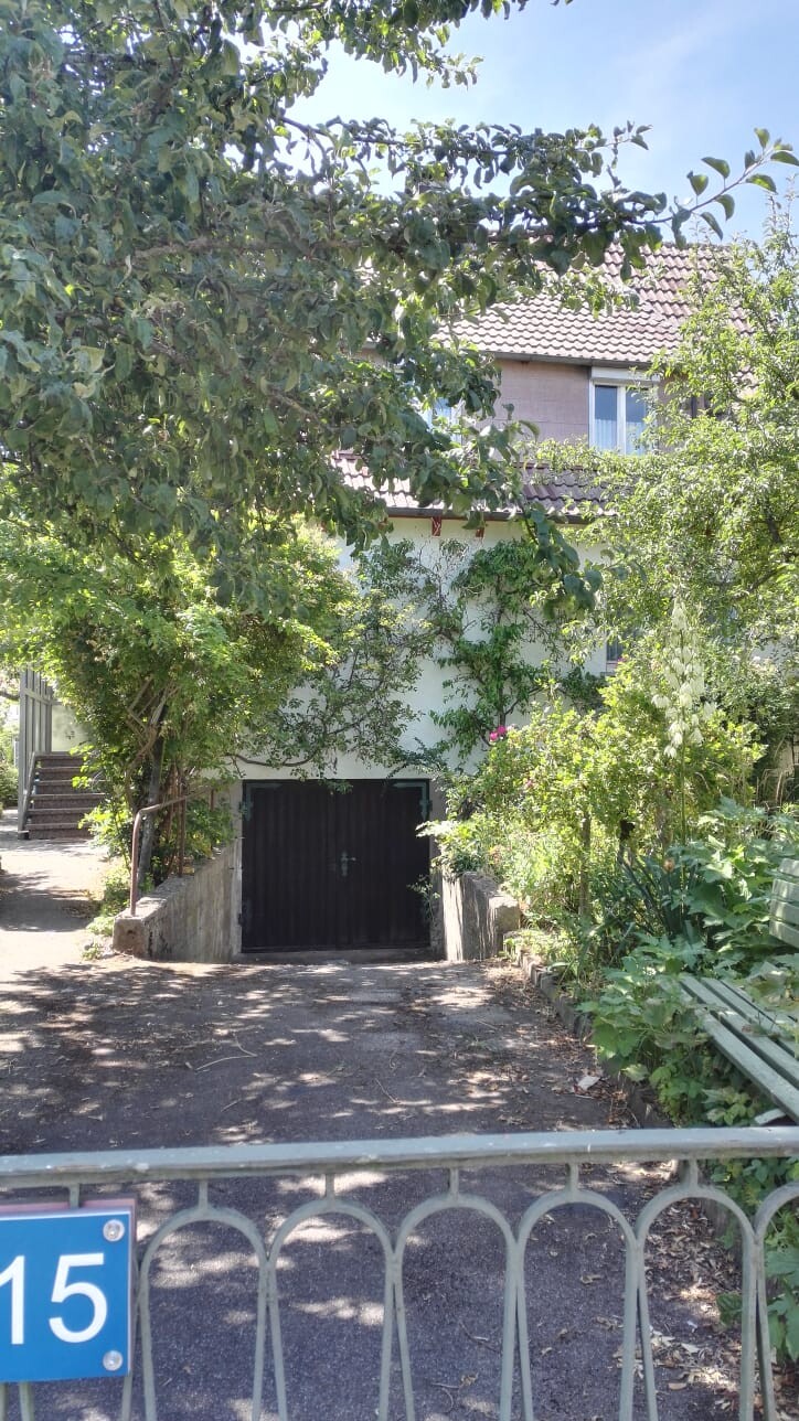 Garageeingang eines Wohnhauses, von Bäumen umgeben; blauer Zaun mit Nummer 15.