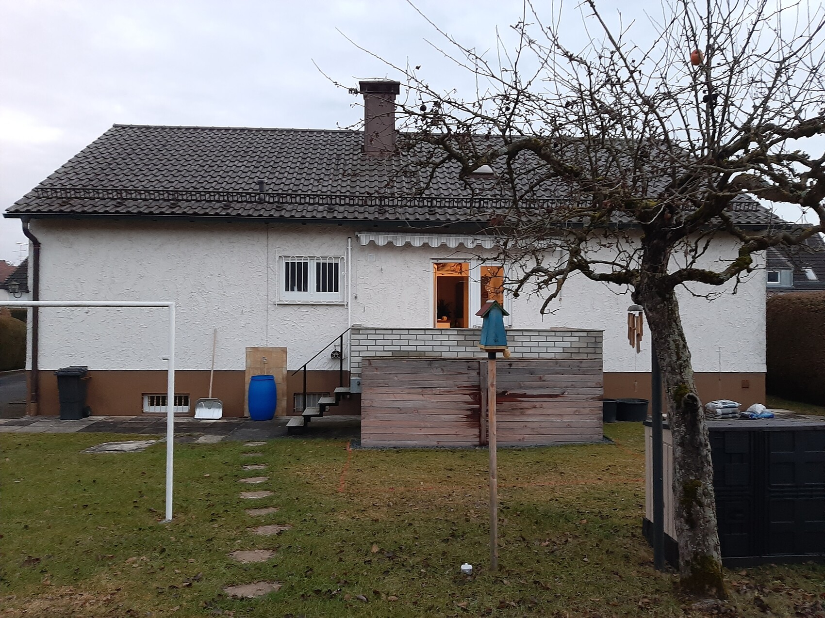 Weißes Einfamilienhaus mit braunem Sockel, Holzterrasse und kahler Baum im Garten.