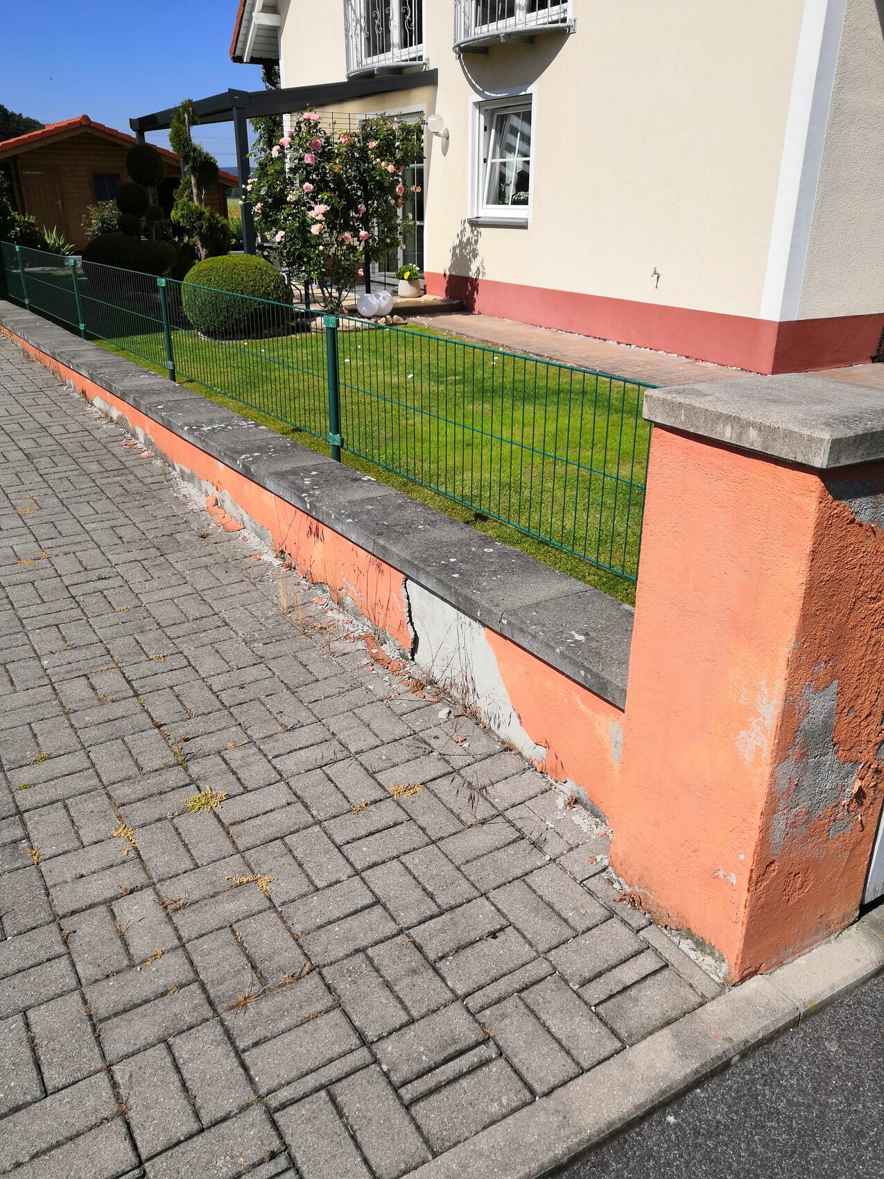 Vorderansicht eines Hauses mit grünem Metallzaun, gepflegtem Garten und Rosenstrauch