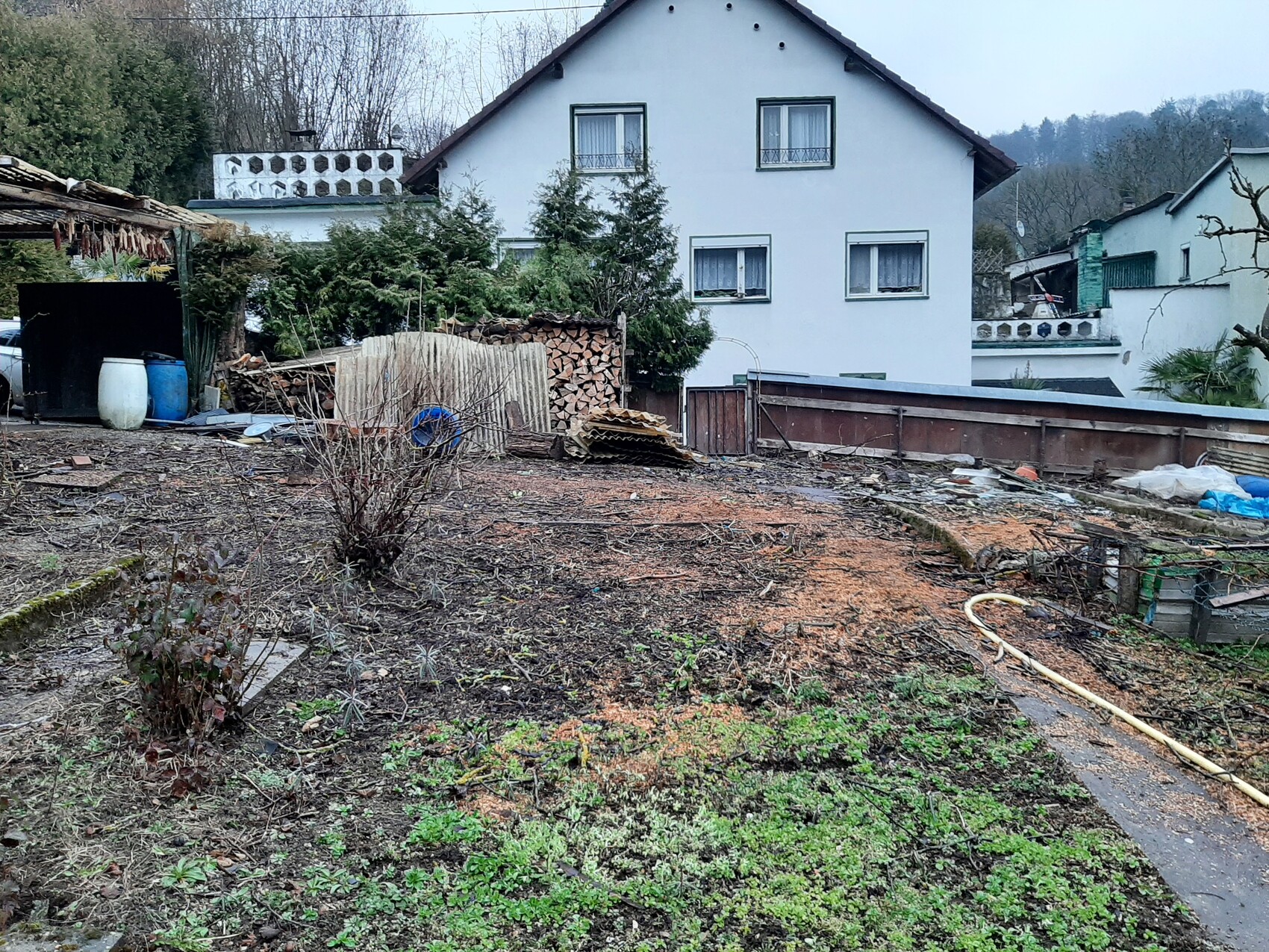 Garten vor einem weißen, zweistöckigen Haus mit Holzstapeln, Schlauch und Bauwerkstoffen.