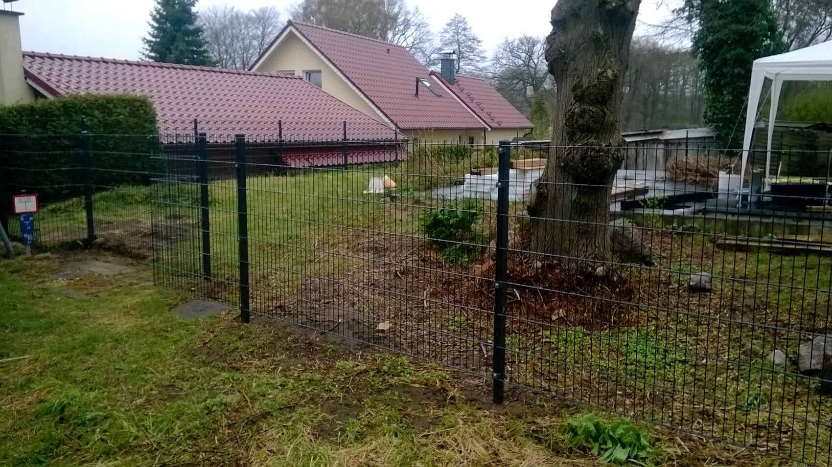 Gitterzaun im Vordergrund, großer Baumstamm rechts, Häuser mit roten Ziegeldächern im Hintergrund.