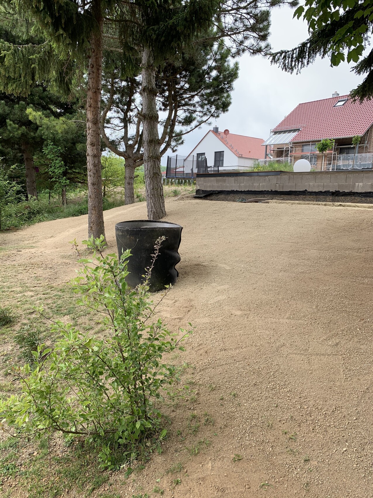 Gartenweg mit Sandboden, Bäumen und großem schwarzen Pflanzkübel; Häuser mit roten Dächern.