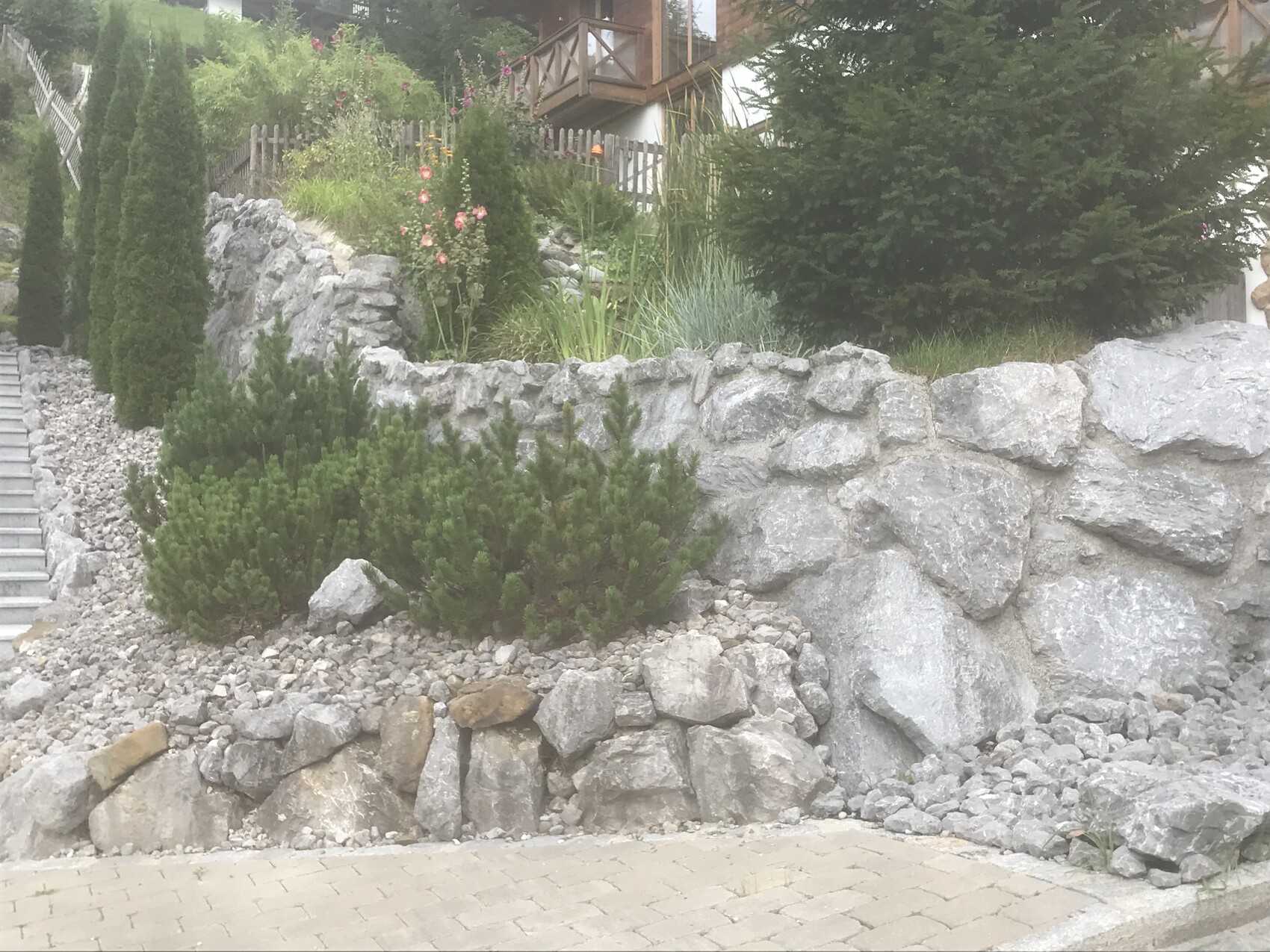 Außengarten mit Steinmauer, Kiesboden und grünem Bewuchs aus Sträuchern.