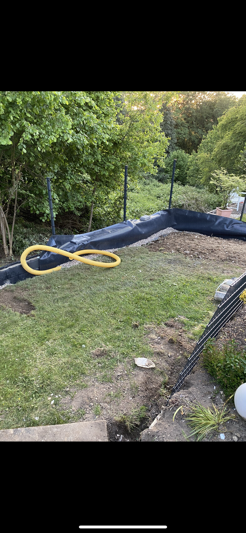 Garten mit ausgehobenem Becken, schwarzer Teichfolien-Liner, gelber Schlauch liegt auf dem Rasen.