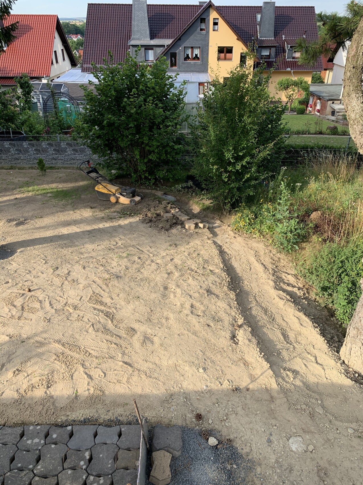 Gartenbaustelle: Sandboden, Rasenmäher, Sträucher und Pflastersteine vor Häusern im Hintergrund