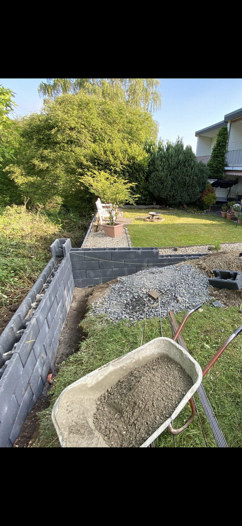 Gartenbaustelle: Betonblockmauer, Schubkarre mit Kies, Kieshaufen, Grünfläche und Bäume.