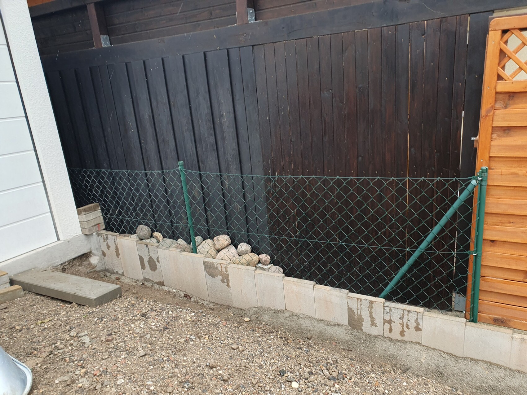 Grüner Maschendrahtzaun vor dunkler Holzwand; Steinblöcke bilden eine Mauer mit Kies davor