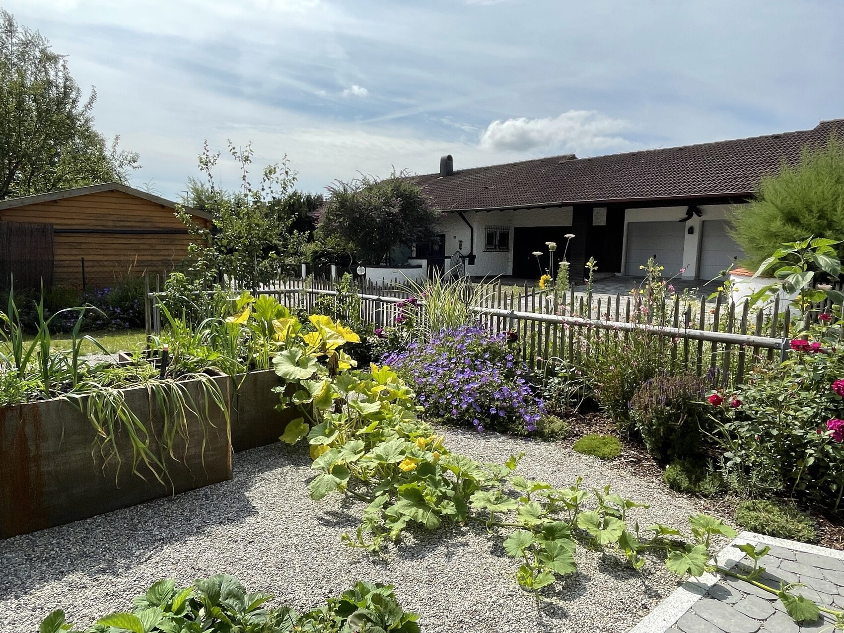 Garten mit Hochbeet aus Holz, bunten Blumen, Kiesweg, Zaun und Haus mit Garage im Hintergrund.