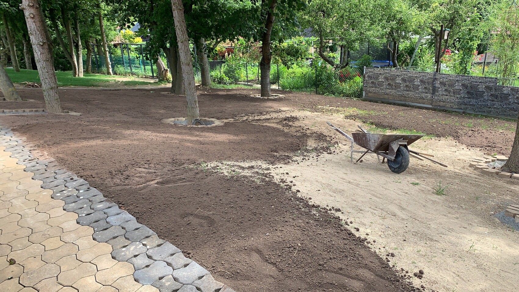 Gartenbereich mit frisch aufgeschüttelter Erde, Schubkarre, Bäumen und linkem sechseckigem Pflasterweg.