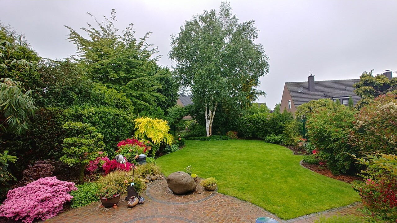 Gepflegter Garten mit grünem Rasen, bunten Blumen, Topfpflanzen und Steinpflasterpfad.