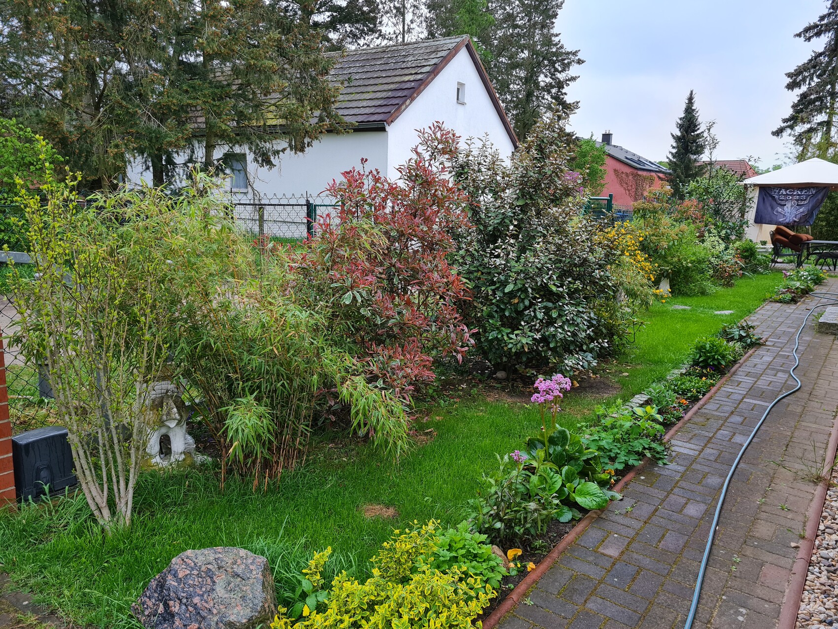 Garten mit Beeten, Sträuchern, Pflasterweg und Blick auf ein Haus im Hintergrund.