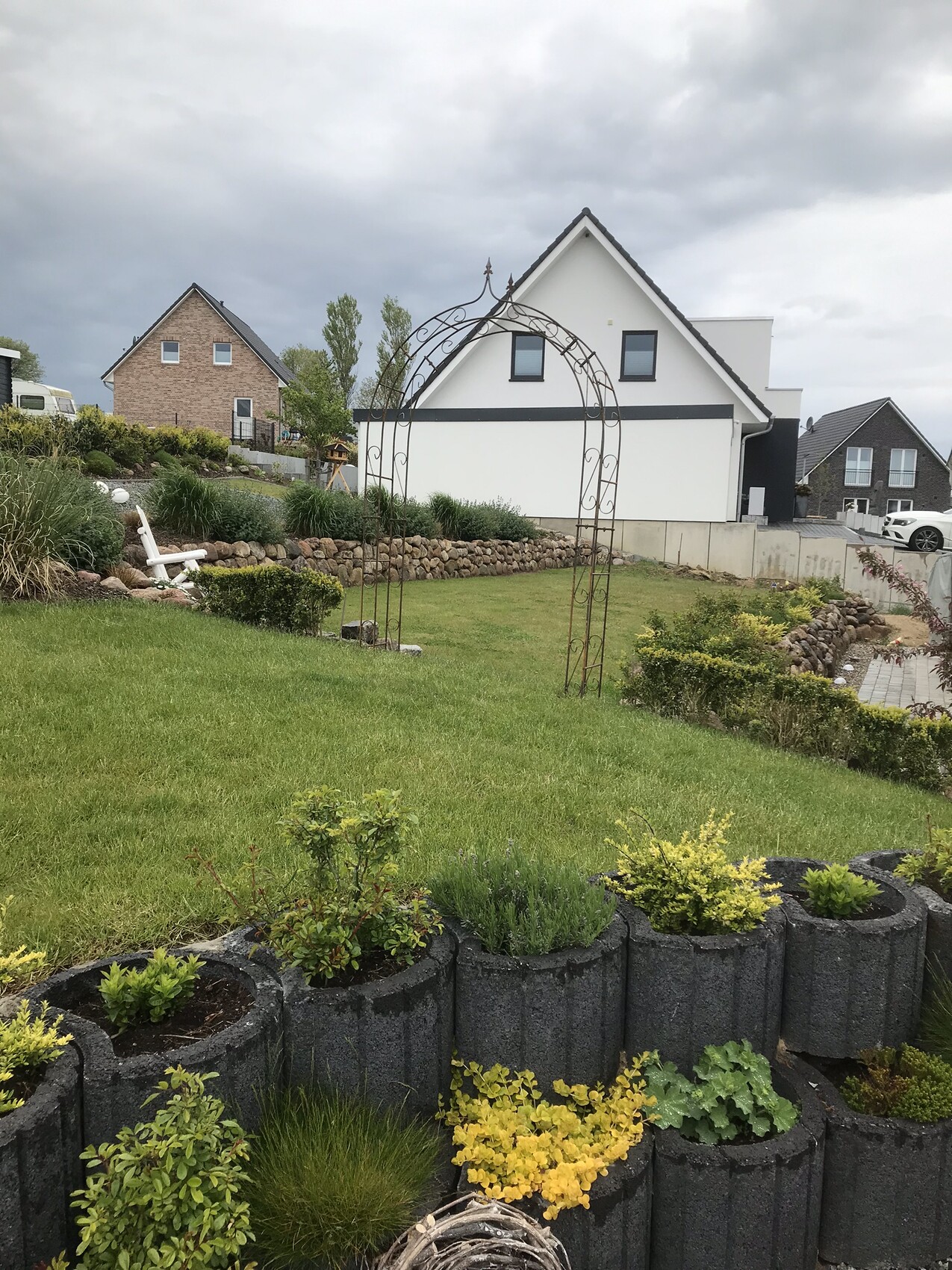 Garten mit grünem Rasen, Steinmauer, Metallbogen-Tor und Häuser im Hintergrund.
