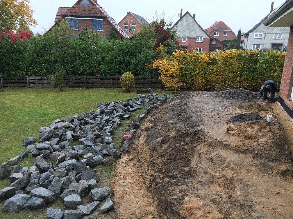 Gartenbaustelle: Großer Erdaushub neben Haus; links Steinblockreihe, rechts Arbeiter.