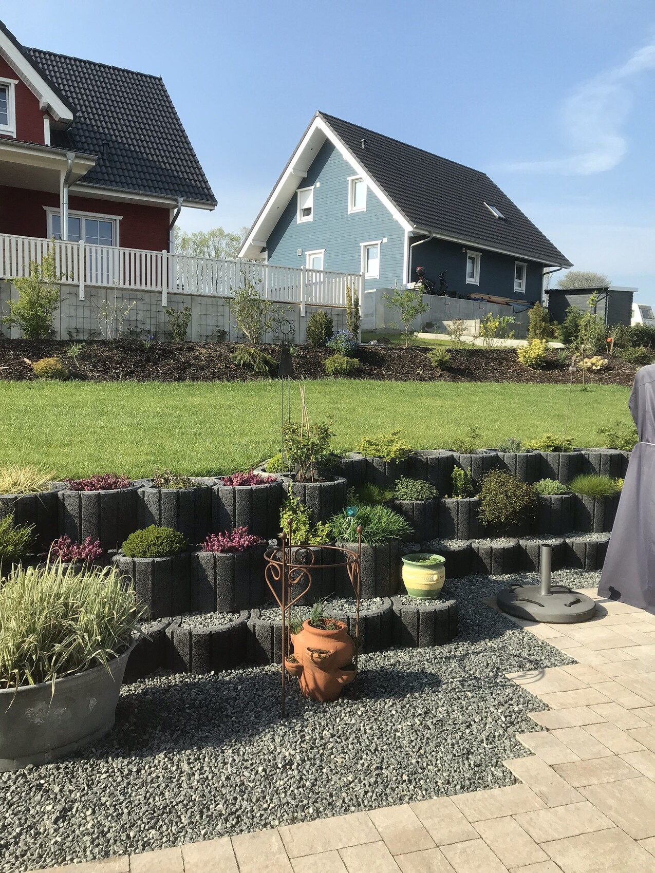 Garten mit Terrasse, Blockstufen, Topfpflanzen und blauem Haus im Hintergrund.