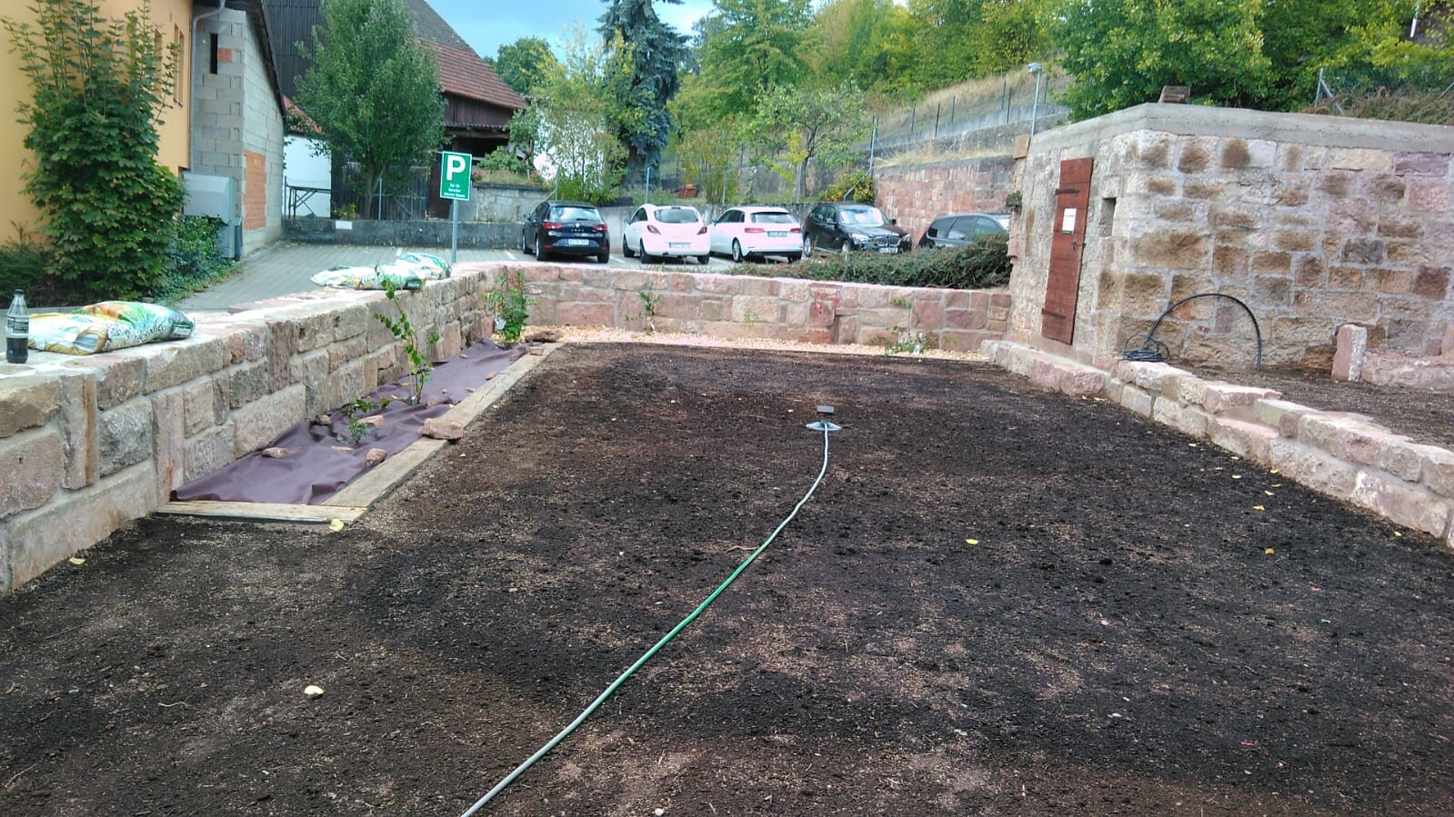 Gartenbaustelle: dunkle Erdfläche, Steinmauer, Schlauch mit Sprinkler; hinter Parkplätze mit Autos.