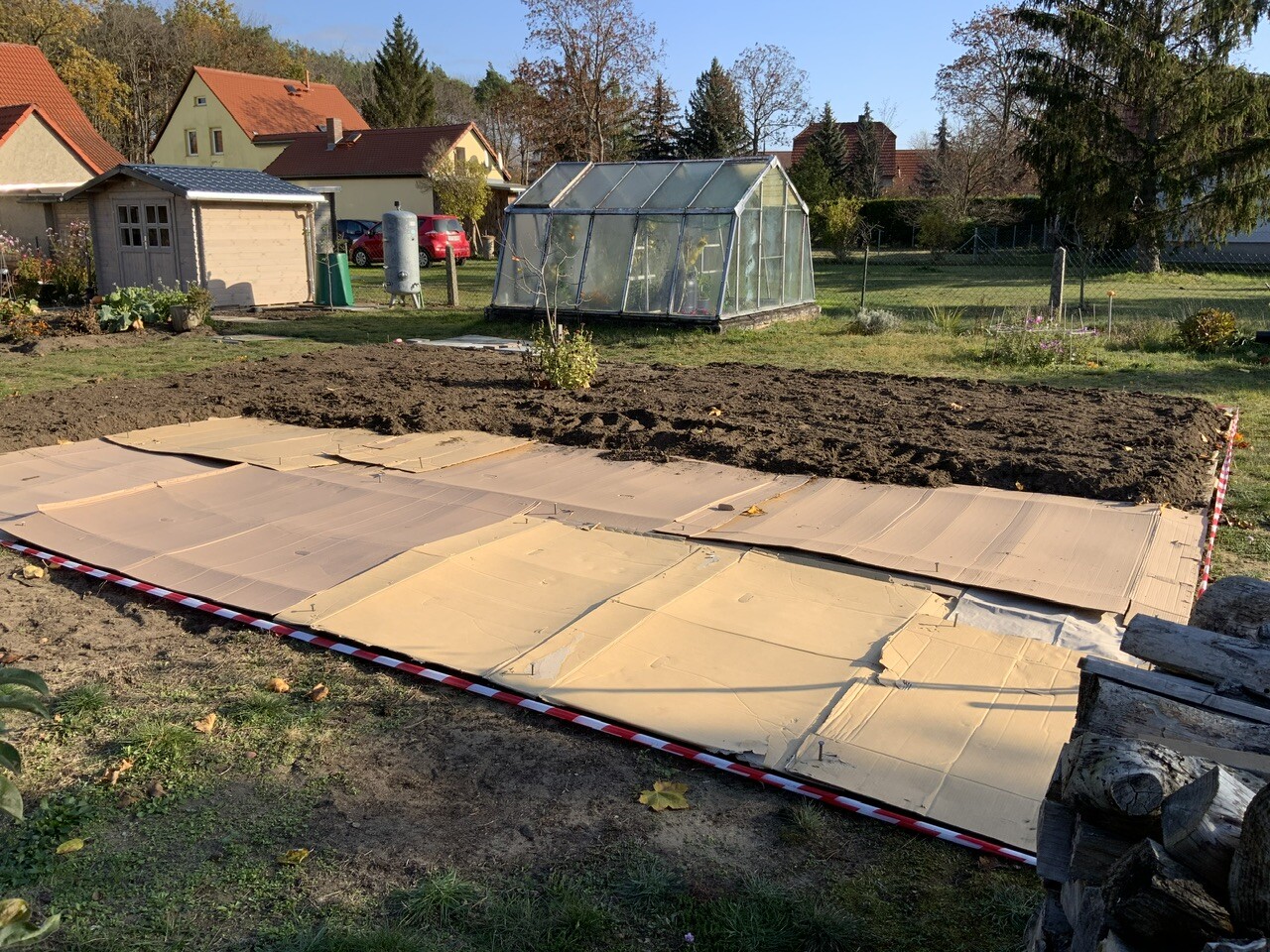Garten mit Gewächshaus, Holzschuppen und Erde unter Abdeckfolie; im Hintergrund Häuser.